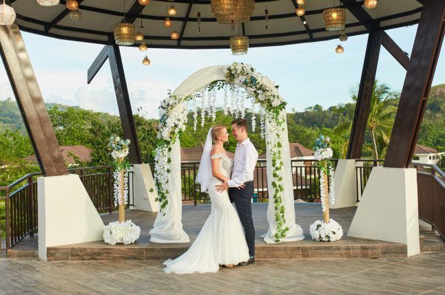 Wedding couple standing under floral arch on an outdoor gazebo, holding hands and kissing.