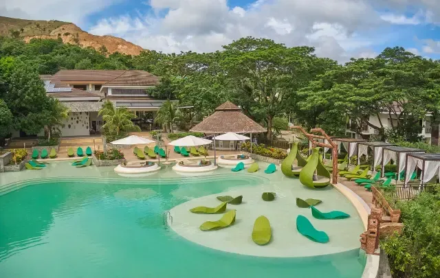 Tropical resort pool with turquoise water, lily-pad islands, palm trees, and lounge chairs beneath cloudy skies