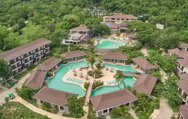 Aerial view of a tropical resort with multiple turquoise pools, brown-roofed buildings, and lush green hills.