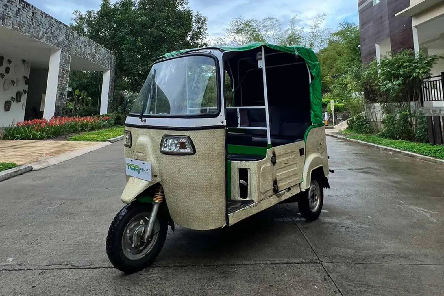 Three-wheeled electric tuk-tuk parked on a wet driveway by buildings and trees.