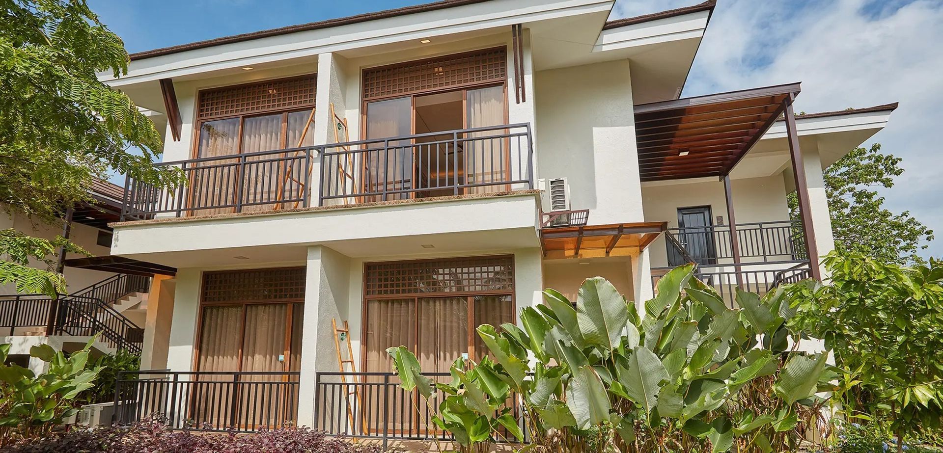 Two-story white house with wooden balconies and lush green plants in front