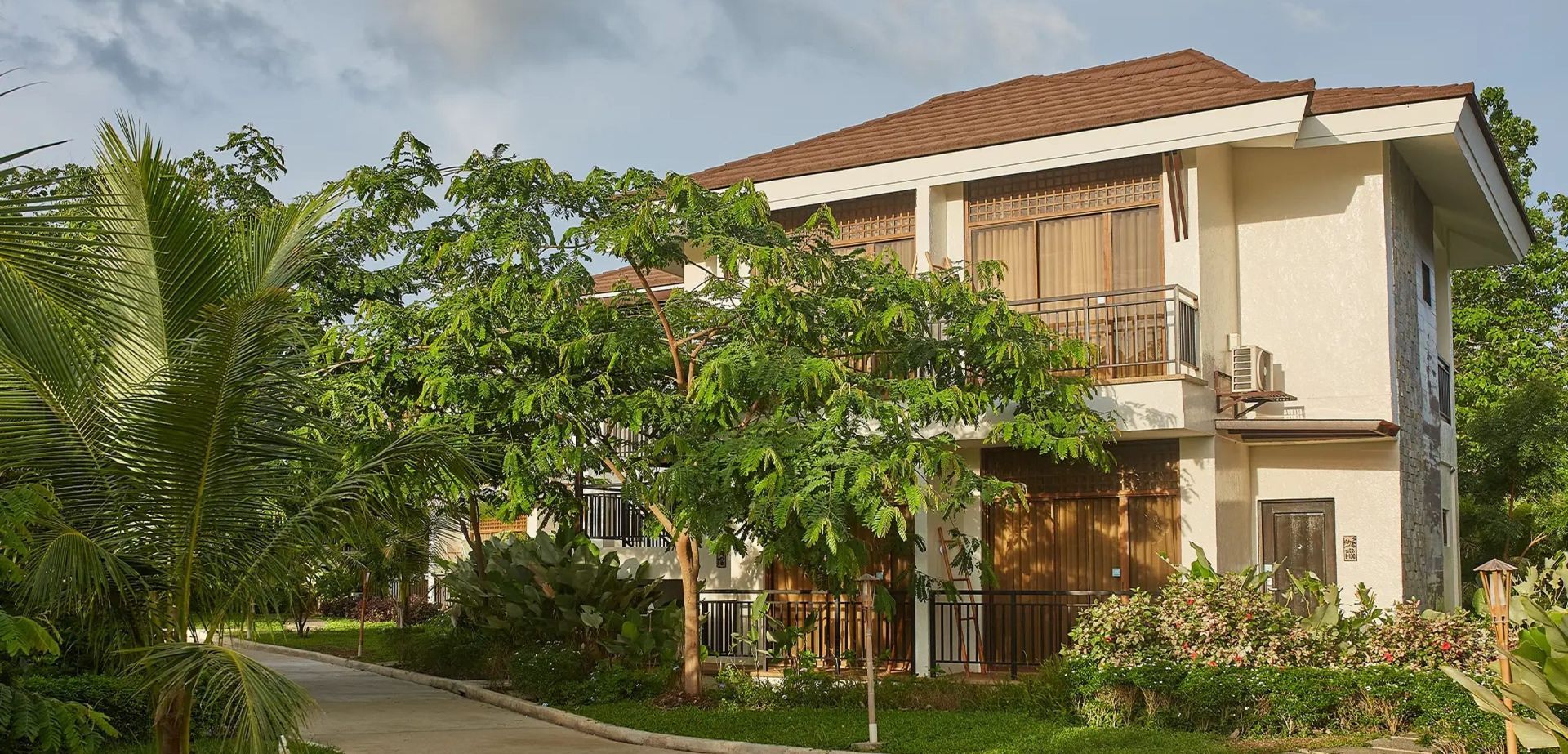 Two-story cream house with red roof amid lush green trees and a garden path