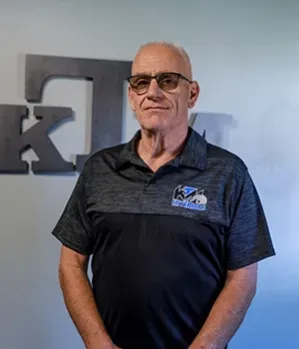 Man with glasses wearing a polo shirt in front of a metal logo.