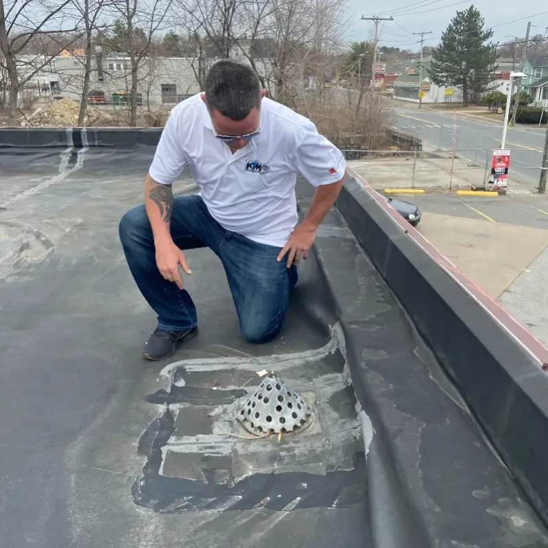 Man on a black rooftop inspecting a drain. He's wearing a white shirt and jeans. Buildings and a road are in the background.