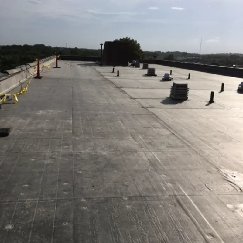Flat, gray commercial rooftop with vents, looking towards a distant tree and buildings, under a clear sky.