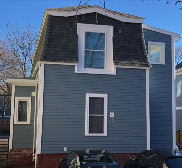 Blue-sided house with a dark roof and white-framed windows. Cars are parked in front of the building.
