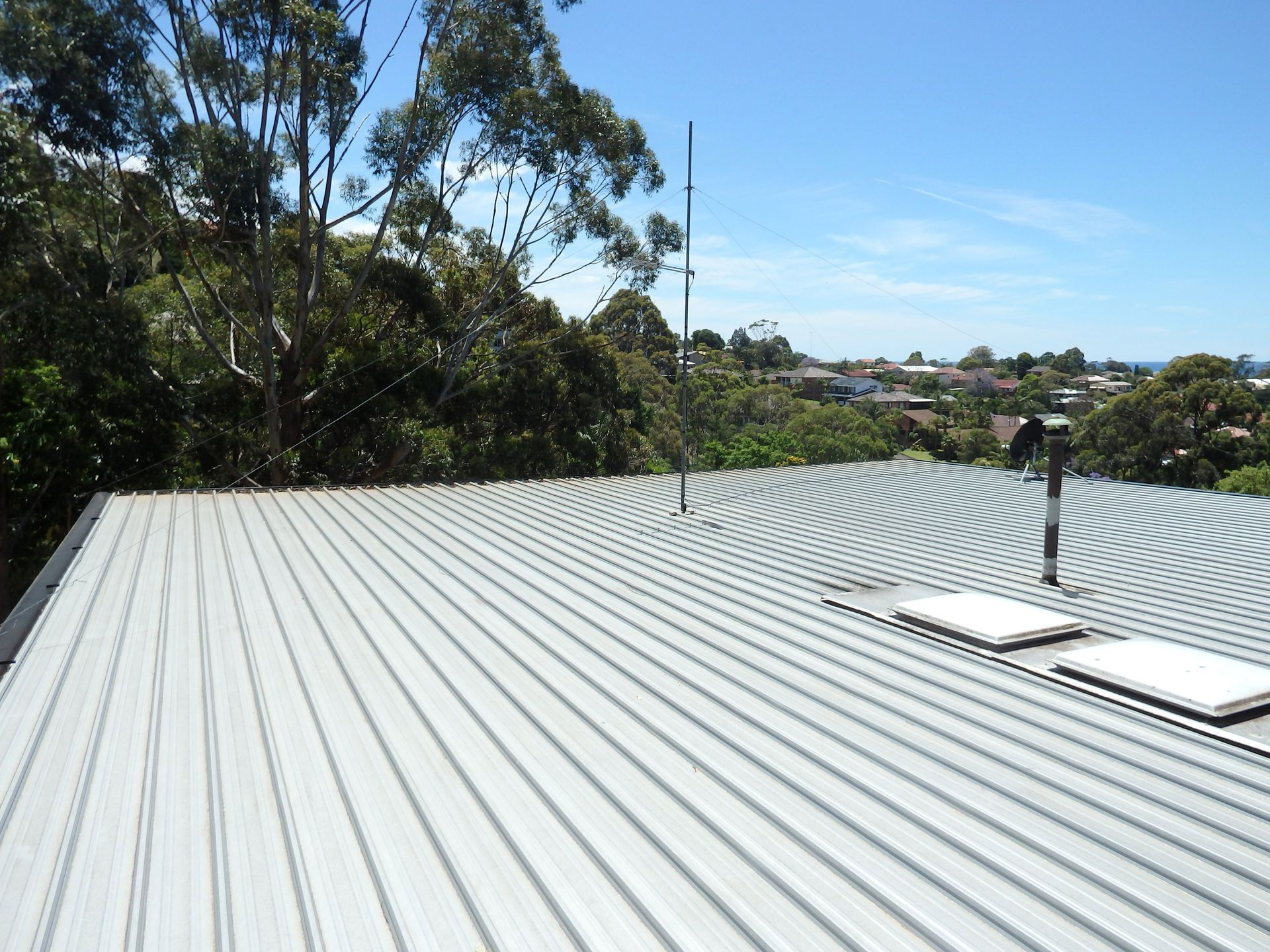 Metal roof with ridged pattern, skylights, and antenna, overlooking trees and a coastal town on a sunny day.