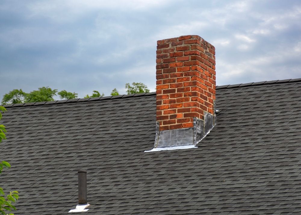 Brick chimney on a dark gray shingle roof, cloudy sky in background.