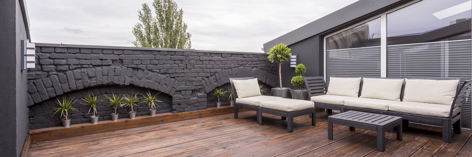 Outdoor patio with gray furniture, black brick wall, and potted plants. Wooden deck under an overcast sky.