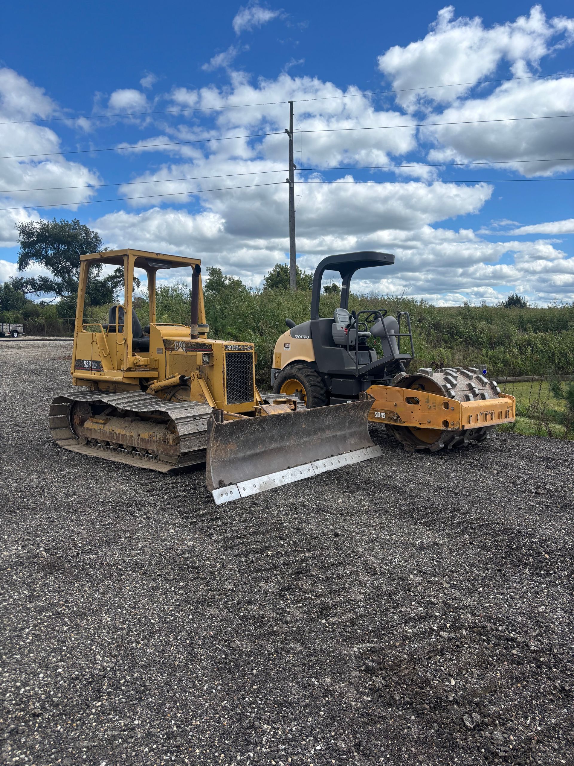 Yellow bulldozer and compact roller parked on gravel under a cloudy blue sky.