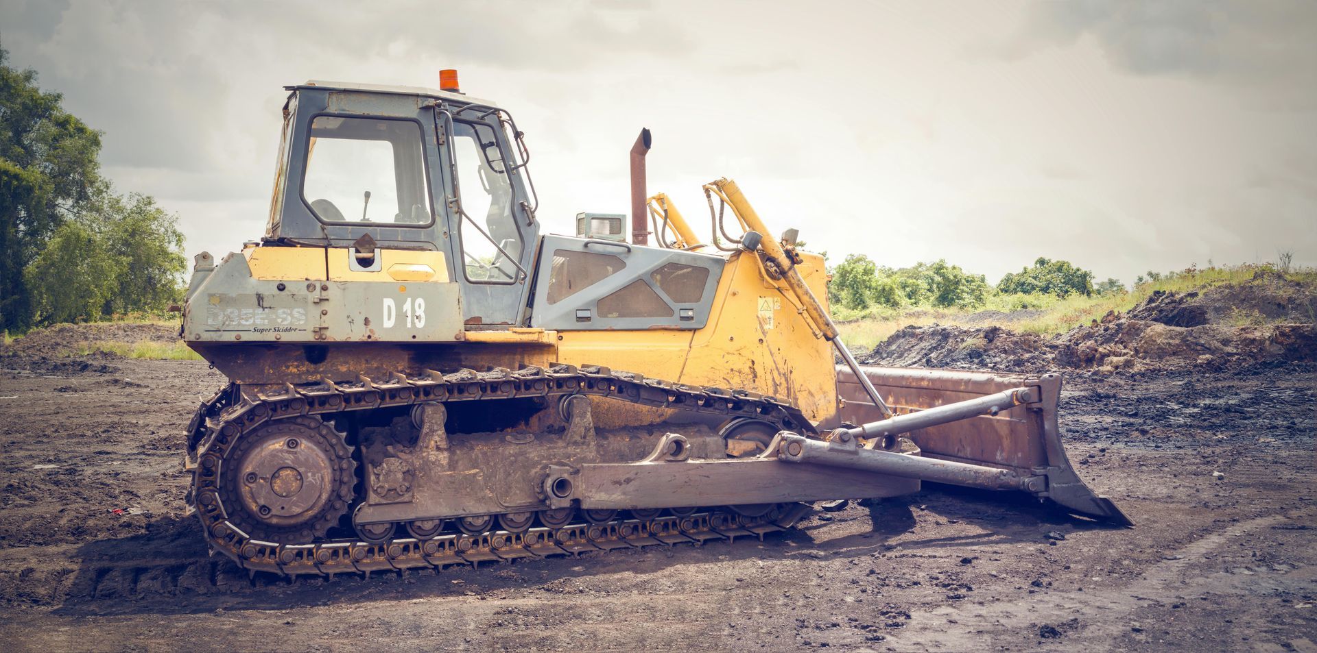 A yellow bulldozer on muddy ground, with trees in the background under an overcast sky.