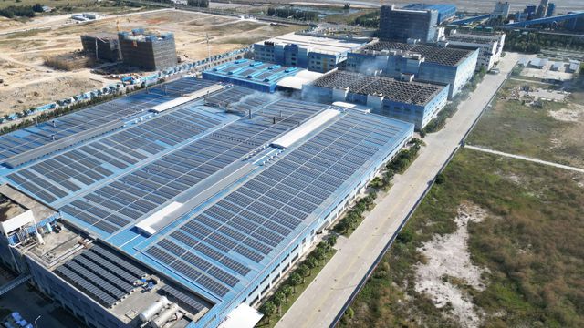 Aerial view of a large industrial solar panel facility beside a road and open land.