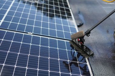 Person pressure-washing blue solar panels on a wet roof