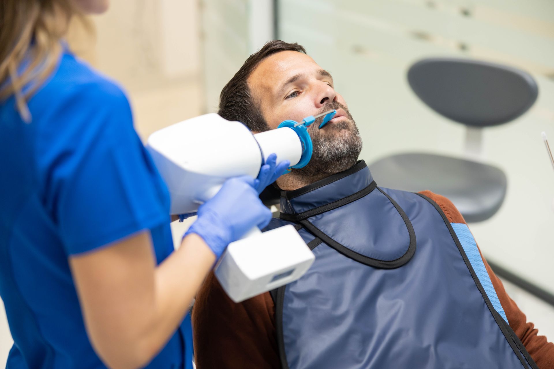 A dentist using a dental instrument on a seated patient in a clinic, with a blue bib and gloves.