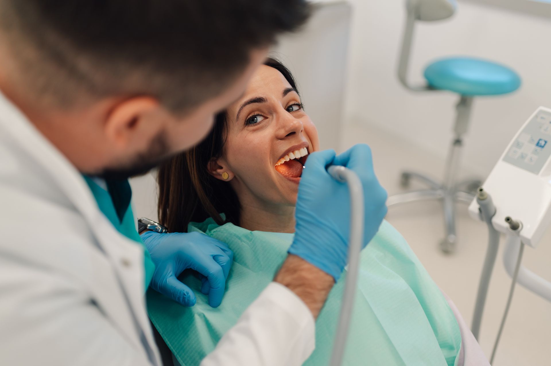 Dentist examining a smiling patient in a dental chair with blue gloves and clinic equipment nearby.