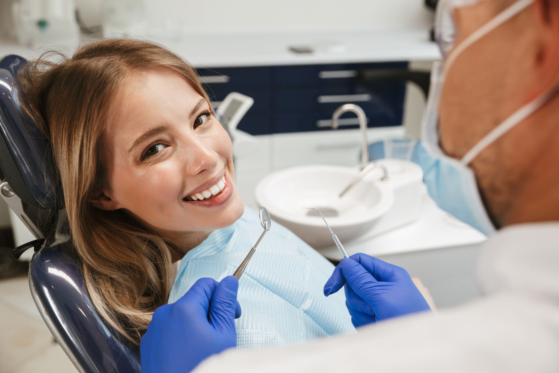 Dentist holding a dental mirror while smiling, a patient sits in a clinic chair.