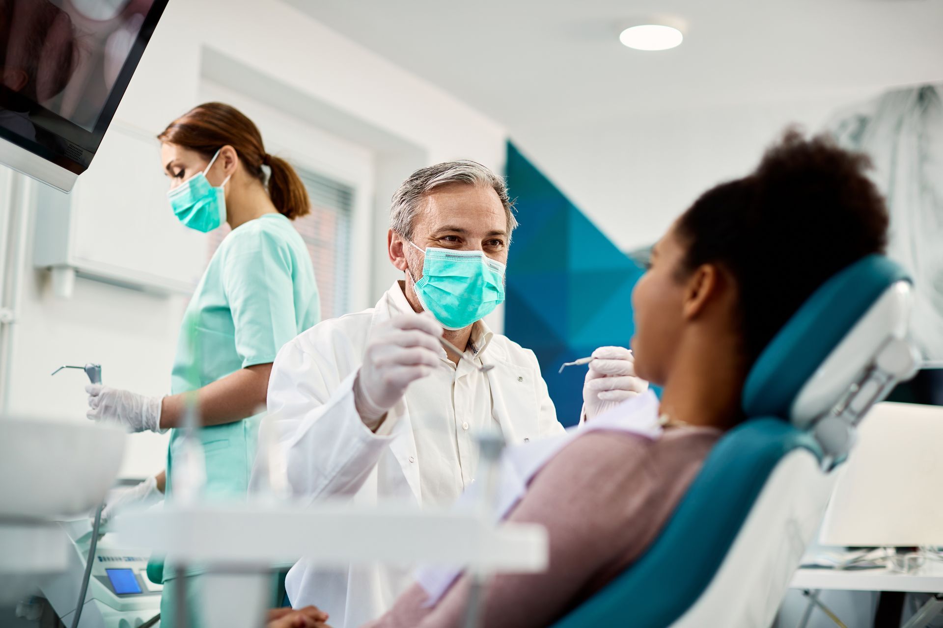Dentist examining a patient in a clinic while a masked assistant stands nearby.