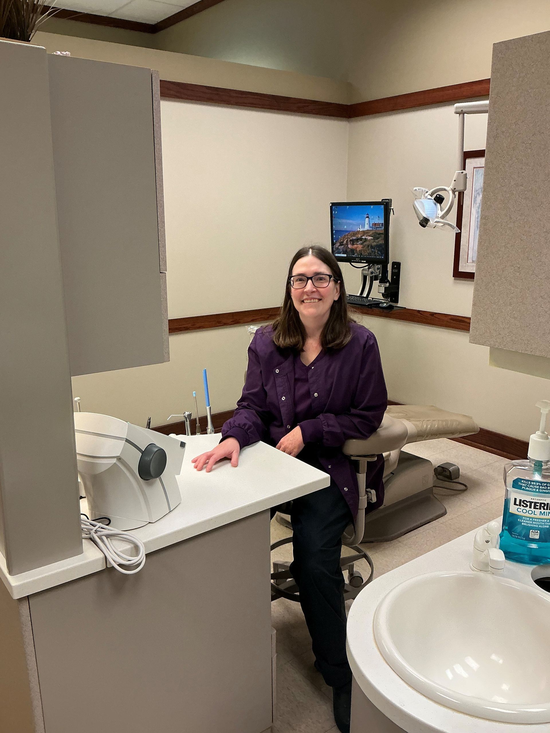 Woman seated at a dental exam chair, smiling in a clinic room beside a sink and counter Woman seated at a dental exam chair, smiling in a clinic room beside a sink and counter