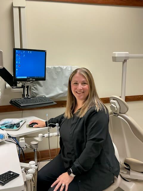 Dentist seated in exam room beside dental chair, smiling at camera with computer monitor in background Dentist seated in exam room beside dental chair, smiling at camera with computer monitor in background