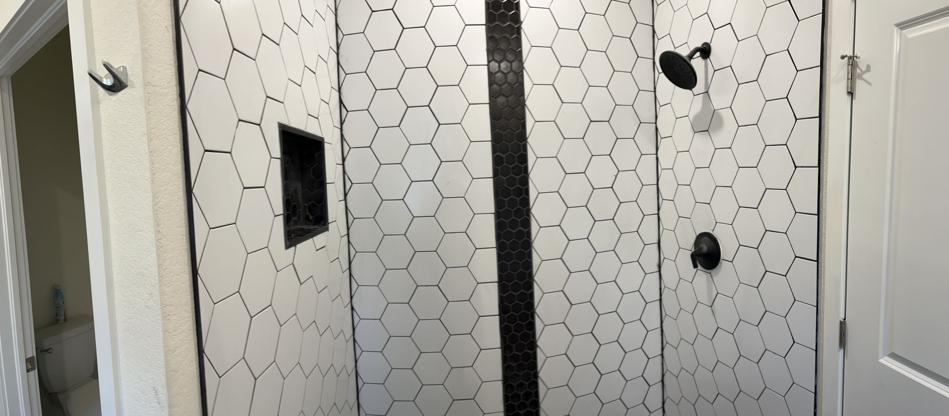 Bathroom shower with white hex tile, black accents, and a glass door; small recessed shelf and shower fixtures visible