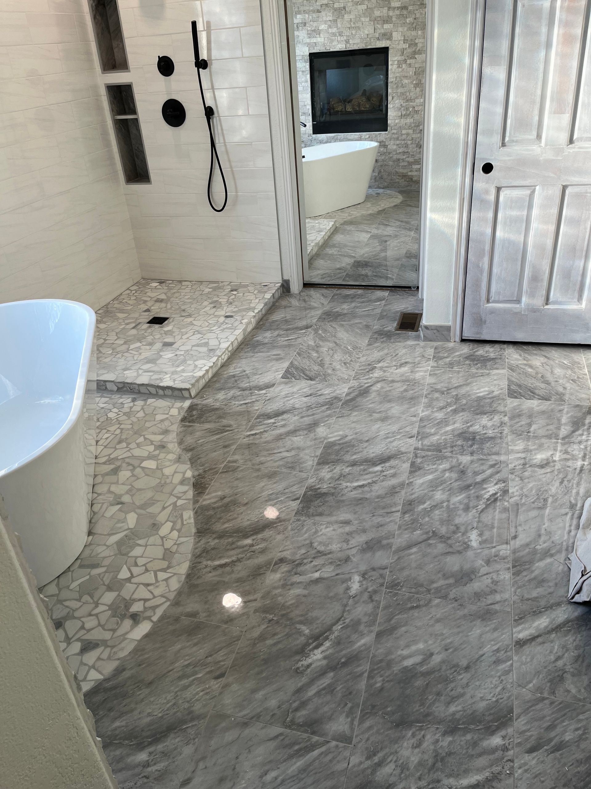 Bathroom with white tub, tiled shower, and large gray floor tiles reflected in a mirror.