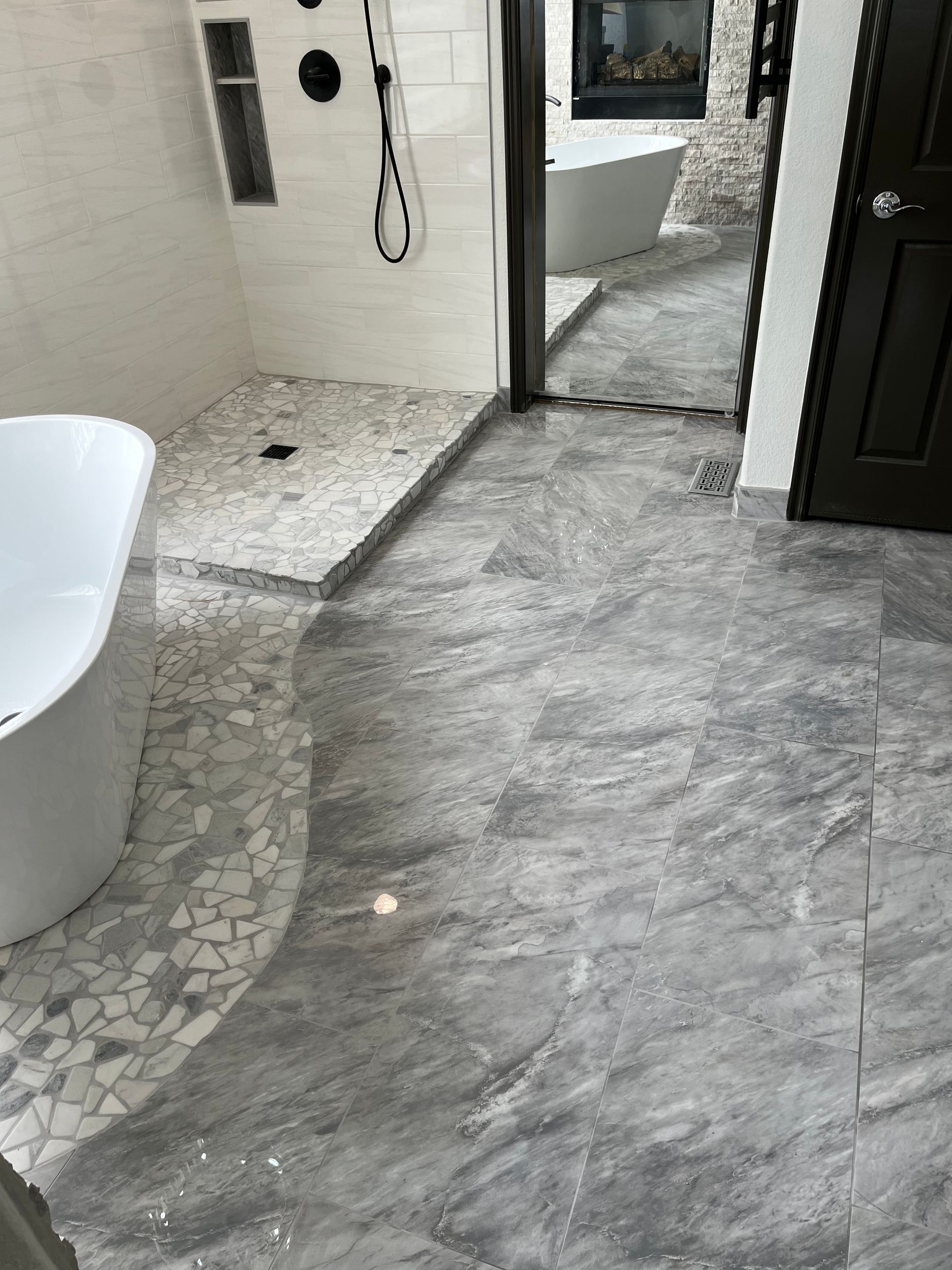 Small bathroom with gray stone-look floor, mosaic shower area, white tub, and open door to another room