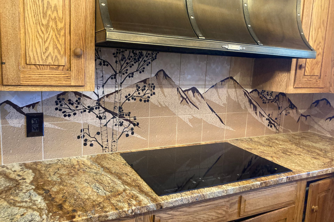 Kitchen with granite countertops, mountain-pattern backsplash, wood cabinets, and a black cooktop under a stainless hood