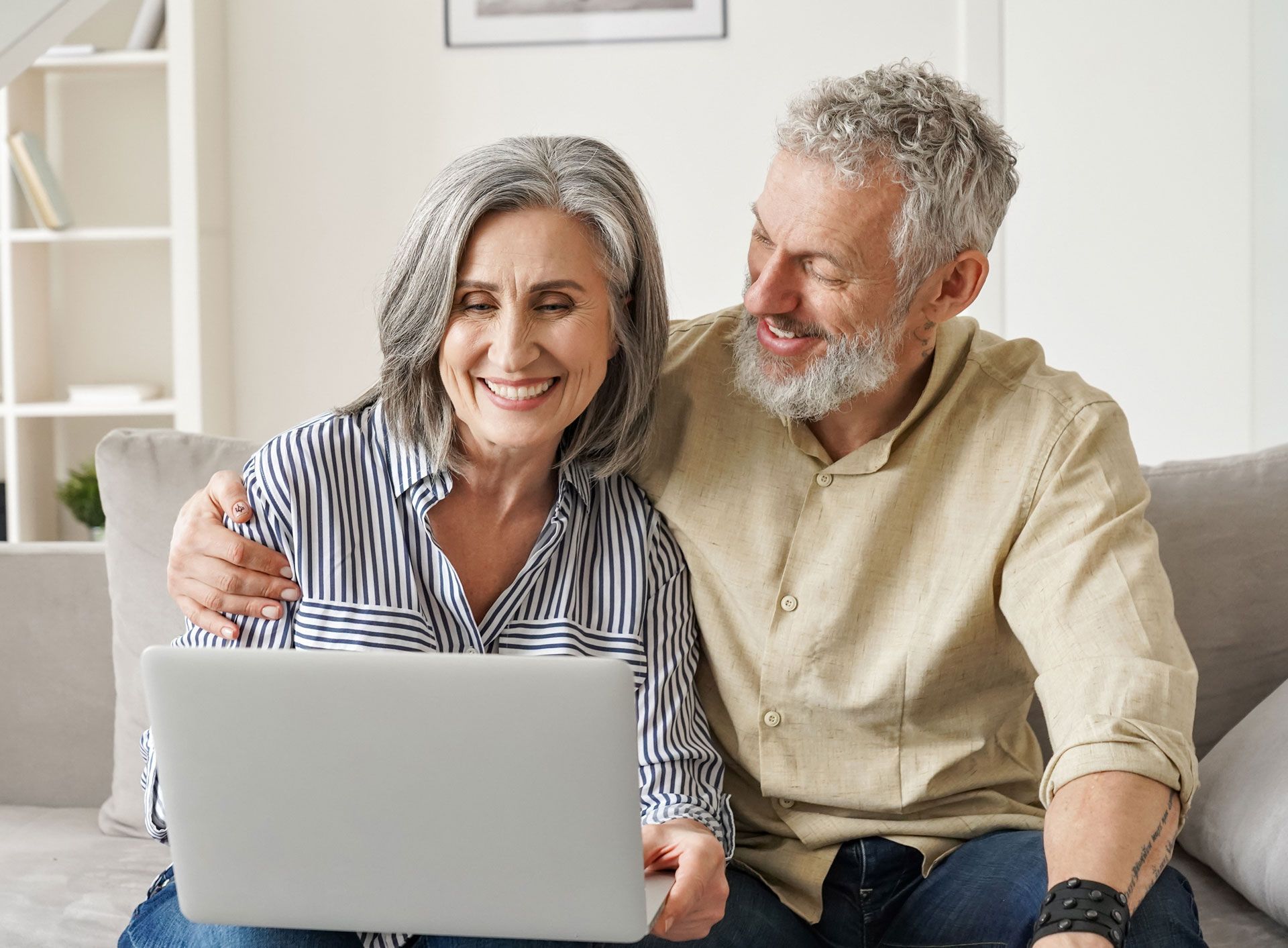 An elderly couple is sitting on a couch using a laptop computer.