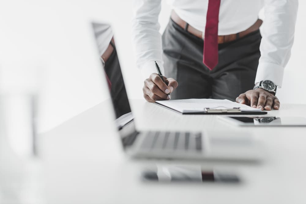 A man in a tie is writing on a clipboard in front of a laptop computer.