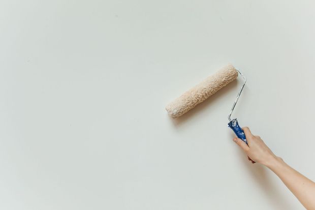 A hand uses a blue-handled paint roller on a white wall.