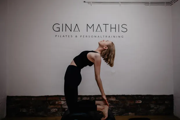 Eine Frau macht in einem Studio eine Rückbeuge vor einer Wand mit der Aufschrift „Gina Mathis“