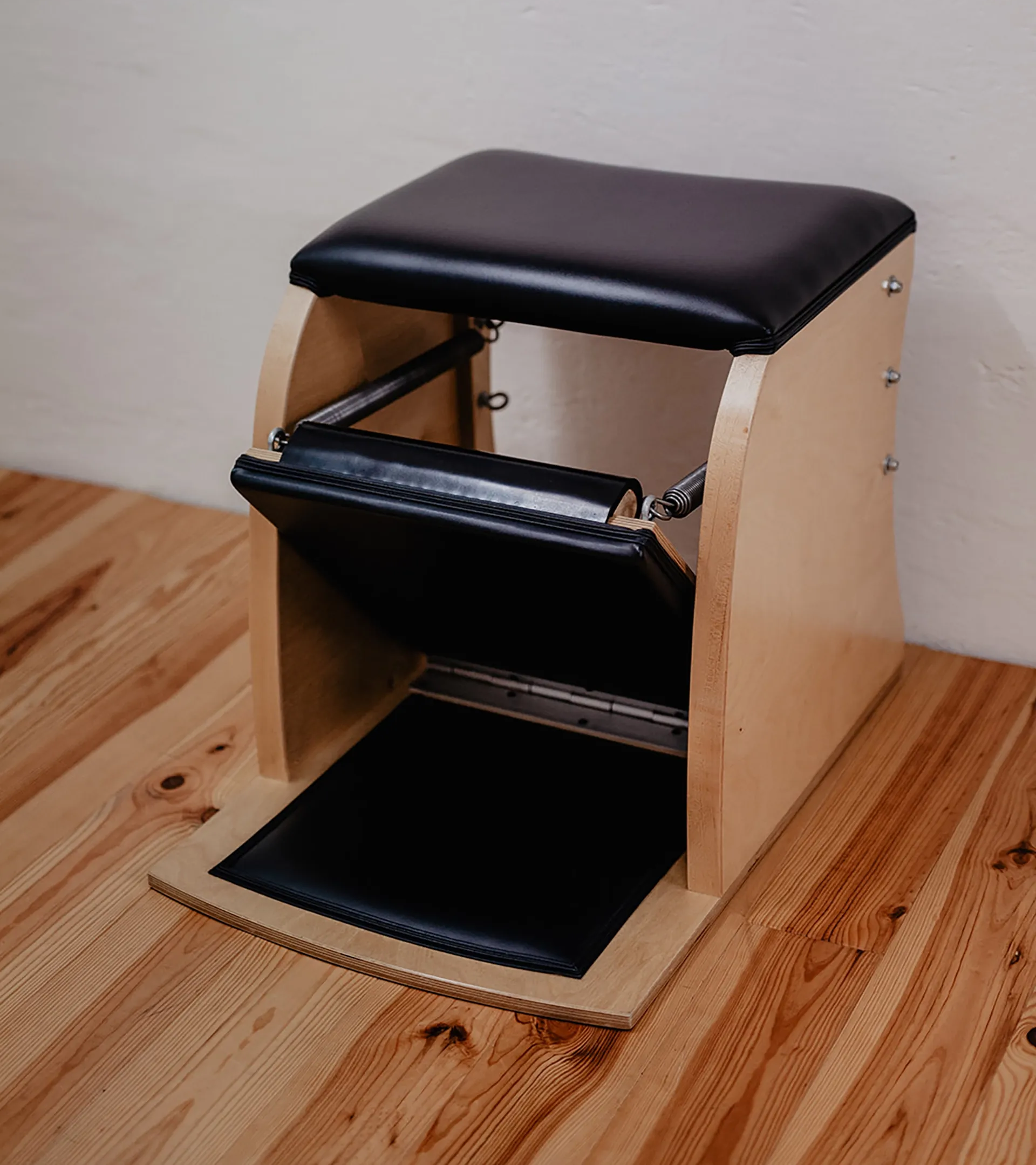 Wunda Chair, ein Holzhocker mit schwarzer gepolsterter Sitzfläche und Ablageboden, steht auf einem Holzboden an einer Wand