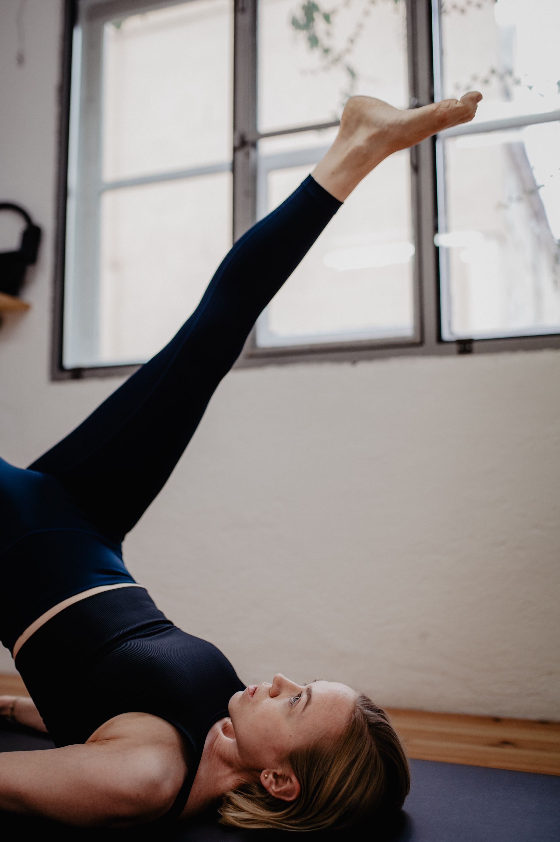 Eine Frau macht drinnen auf einer Matte eine Yoga-Pose und hebt ein Bein in Richtung eines hellen Fensters.