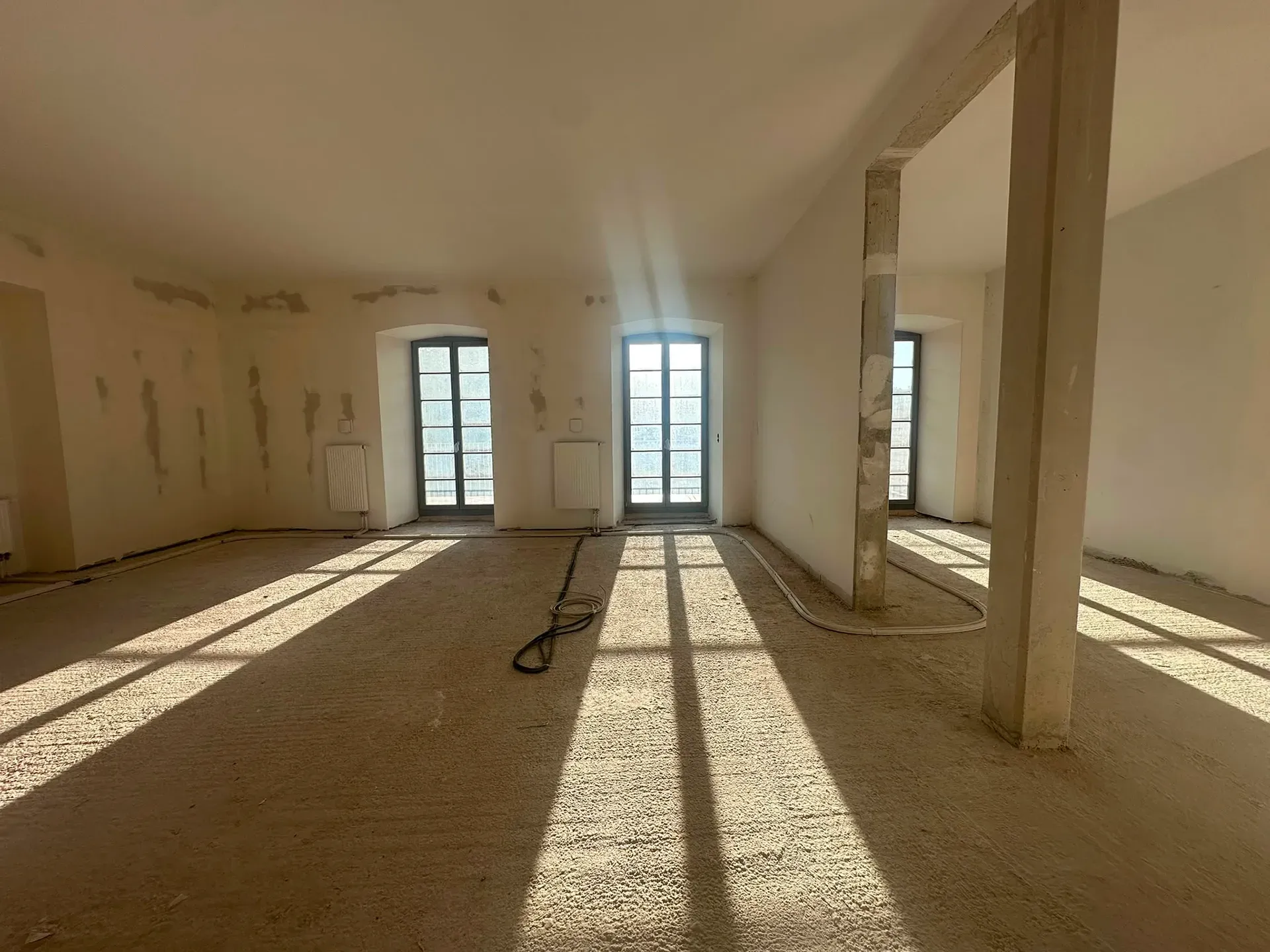 Leerer, unfertiger Raum mit weißen Wänden, Säulen und Sonnenlicht, das durch hohe Fenster auf einen staubigen Boden fällt