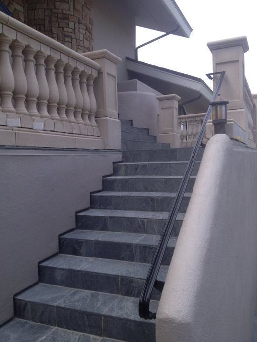Outdoor stone staircase with a black handrail beside a beige house and balustrade.