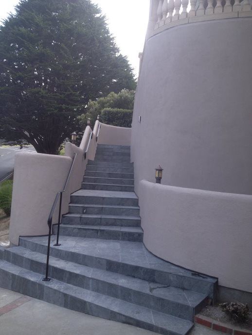 Steep outdoor stone stairs beside a curved beige building, with a black handrail and trees nearby