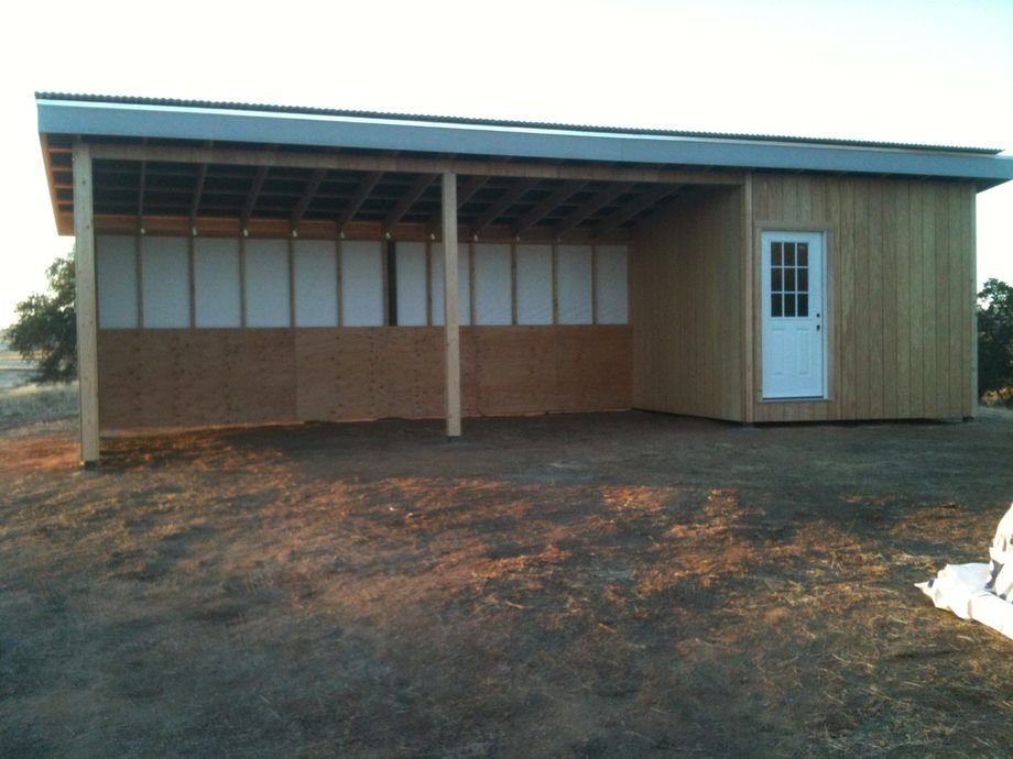 Small unfinished wooden building with a metal roof, white door, and open front on a dirt lot.