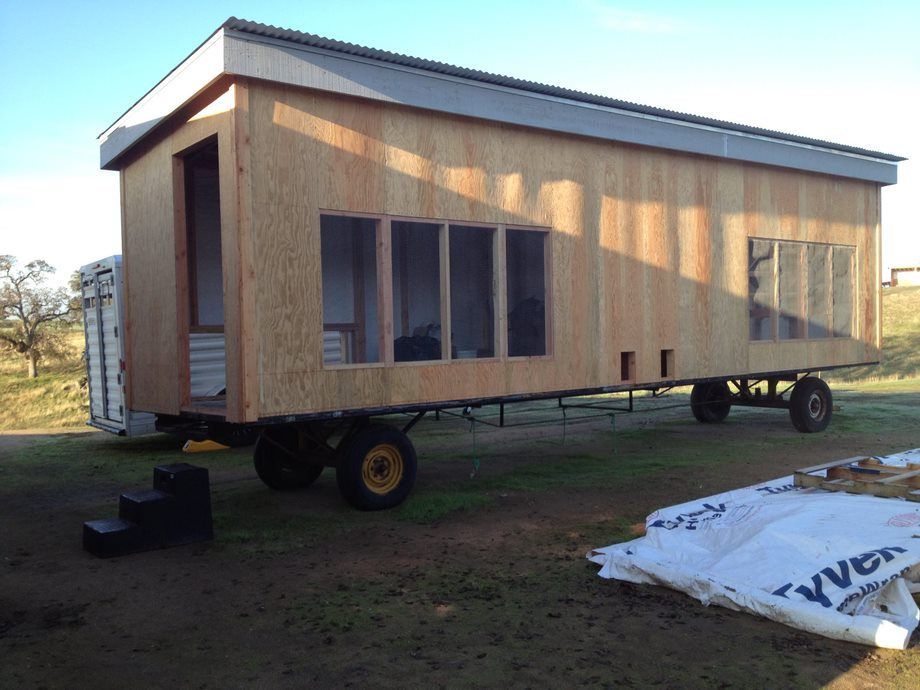 Tiny house on a trailer with wood siding, large windows, and a metal roof in a grassy yard.