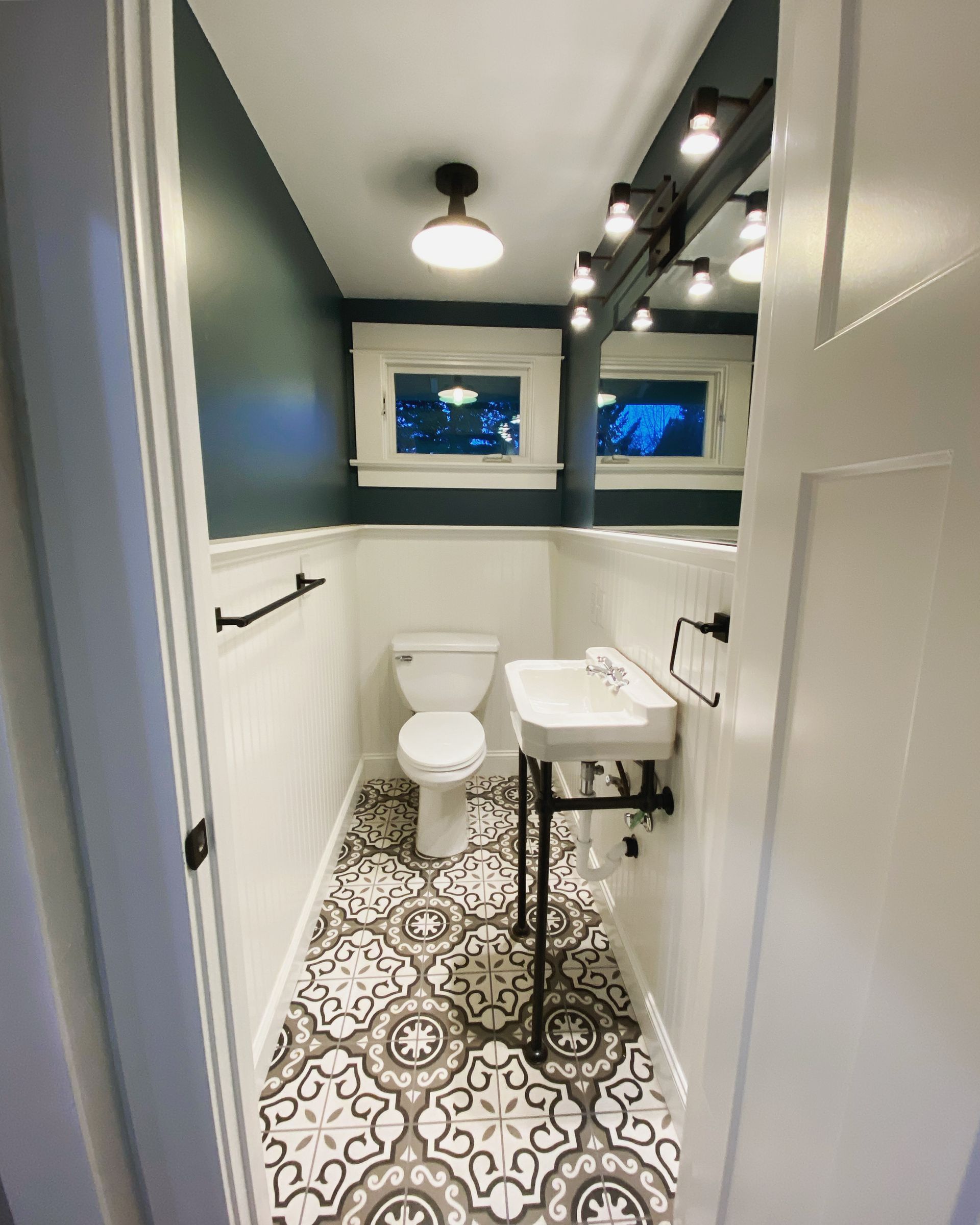 Small modern bathroom with patterned tile floor, white toilet and sink, blue walls, and two small windows.