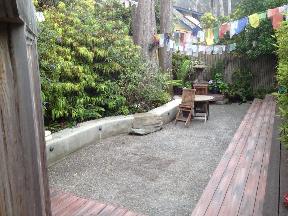 Small patio garden with gravel, wooden benches, chairs, and hanging prayer flags between trees.
