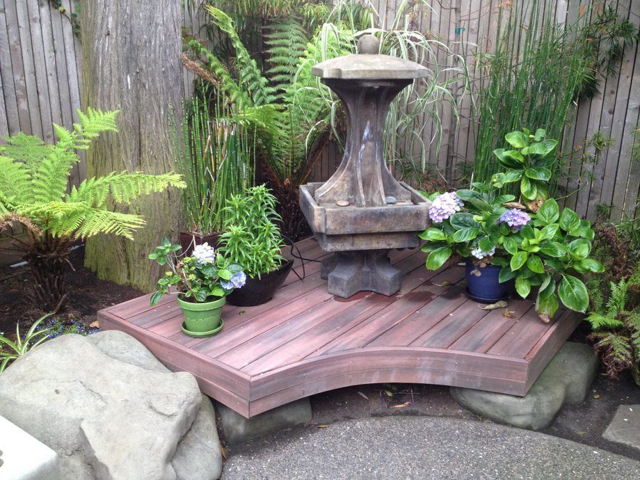 Small backyard garden with a stone fountain on a wooden deck, surrounded by potted ferns and flowers.