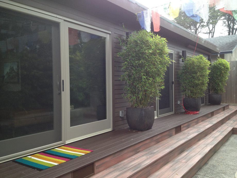 Modern patio with sliding glass doors, wooden deck, potted topiary, and a colorful striped doormat