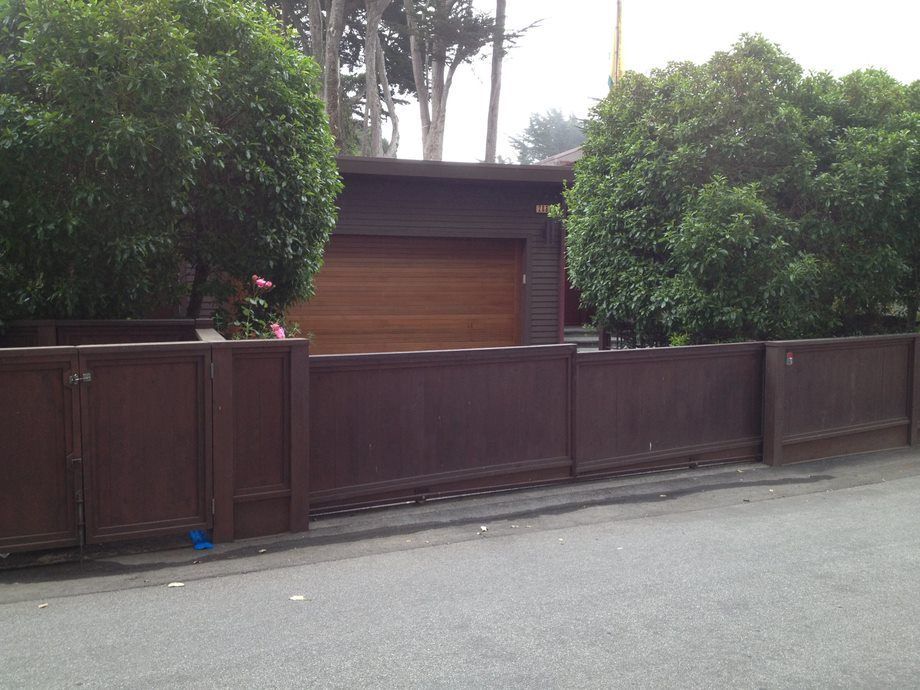 Brown wooden fence and gate in front of a house, with large green trees along the street.