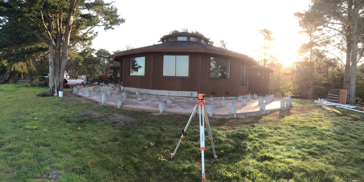 Cabin with a red roof in a grassy yard at sunset, with a survey tripod in the foreground