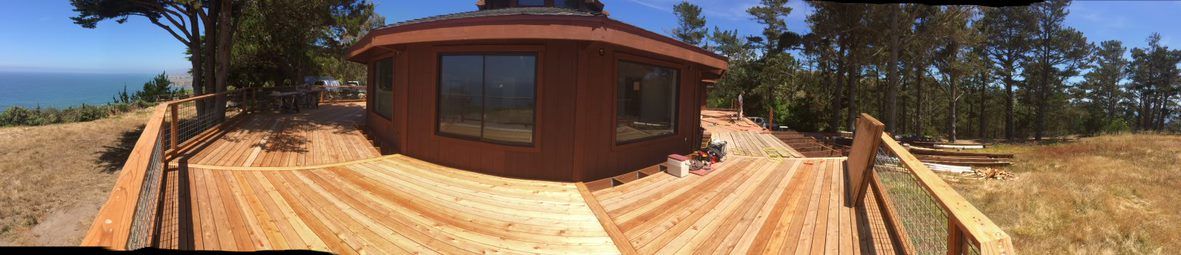 Wide-angle view of a wooden cabin under construction on a dirt lot with trees and blue sky