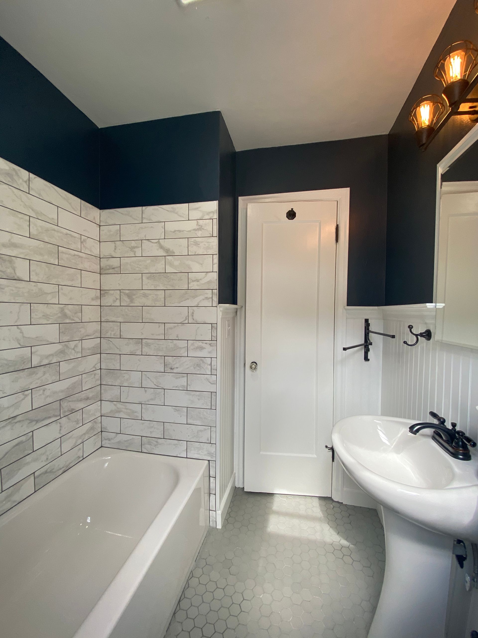 Small bathroom with white tub, sink, door, and navy walls; white tiled tub surround and patterned floor.