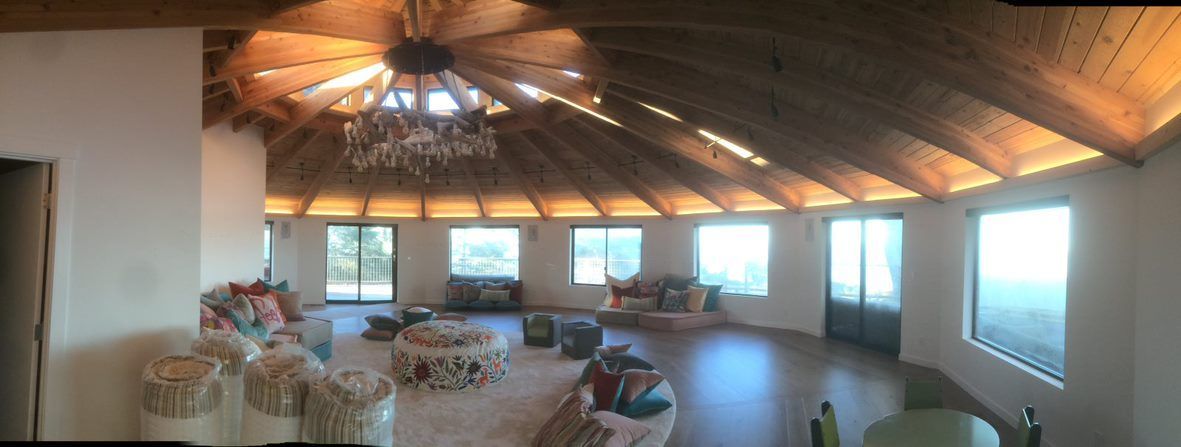 Sunlit rustic attic room with wooden beams, chairs, and large windows overlooking a lake
