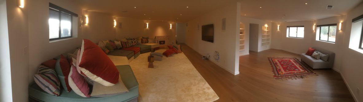 Wide living room with warm lighting, hardwood floors, a sofa with red pillows, and a rug area in the center.