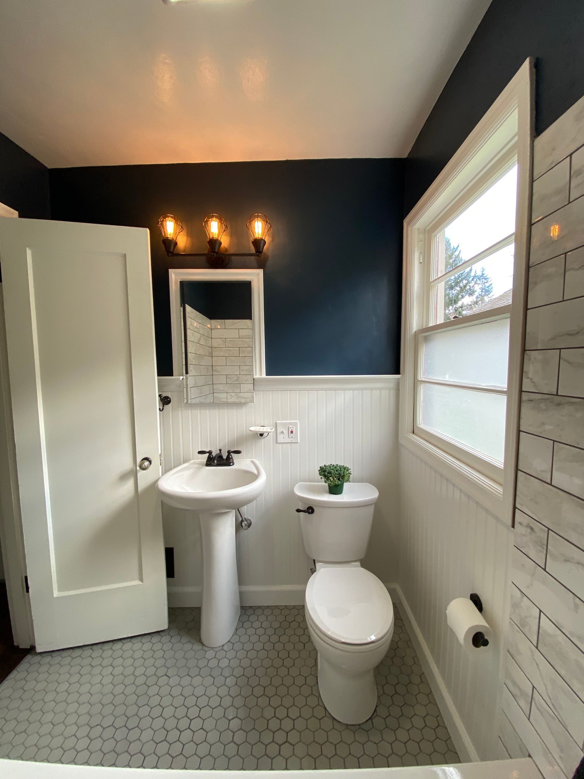 Small bathroom with a pedestal sink, toilet, white-and-navy walls, hex tile floor, and a window.