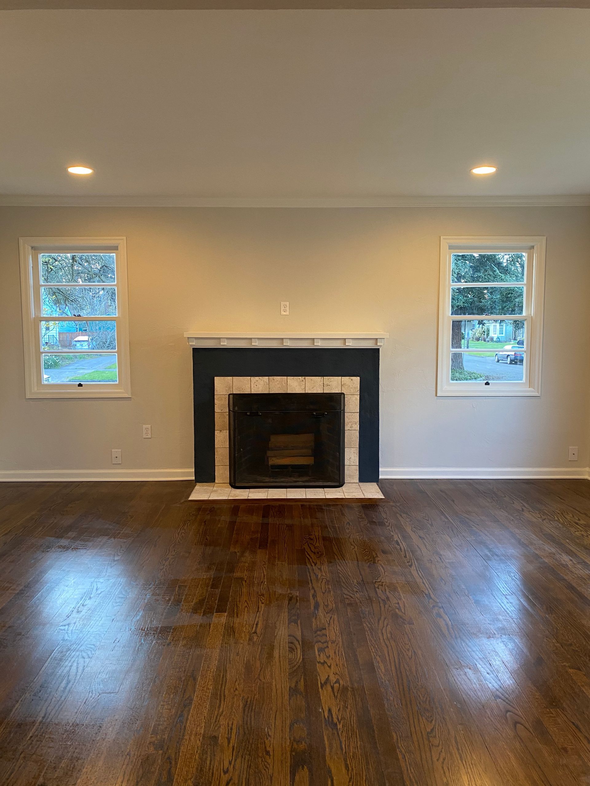 Empty living room with dark hardwood floors, white walls, two windows, and a black fireplace mantel